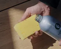 Et nærbillede af en hånd, der holder en rektangulær gul svamp med små porer. En anden hånd hælder væske fra en sølvfarvet metalflaske ned på svampen. Baggrunden er en lys træoverflade, og lyset er naturligt med bløde skygger.