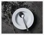 En rund hvid tallerken med diskret rillet kantdesign, placeret på en mørk, tekstureret overflade. En spiseske i rustfrit stål med et enkelt, moderne skaft ligger inde i tallerkenen. Sorte bønner og tørrede urter er kunstnerisk spredt omkring tallerkenen og giver et rustikt udtryk til præsentationen.