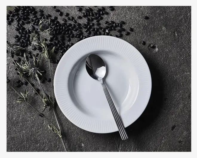 En rund hvid tallerken med diskret rillet kantdesign, placeret på en mørk, tekstureret overflade. En spiseske i rustfrit stål med et enkelt, moderne skaft ligger inde i tallerkenen. Sorte bønner og tørrede urter er kunstnerisk spredt omkring tallerkenen og giver et rustikt udtryk til præsentationen.