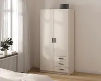 Et højt, fritstående garderobeskab med en glat, mat hvid overflade. Skabet har to store låger med sorte, rektangulære greb og tre rummelige skuffer nedenunder, hver med matchende sorte greb. Garderobeskabet har et rent, moderne design med lige linjer og ingen synlig udsmykning. En rund, grå opbevaringsboks står ovenpå. Skabet står på en flad sokkel og er placeret i et lyst rum med trægulv.