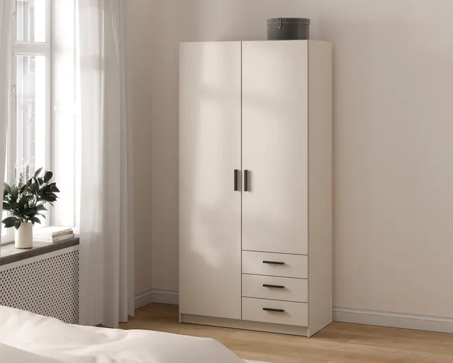 Et højt, fritstående garderobeskab med en glat, mat hvid overflade. Skabet har to store låger med sorte, rektangulære greb og tre rummelige skuffer nedenunder, hver med matchende sorte greb. Garderobeskabet har et rent, moderne design med lige linjer og ingen synlig udsmykning. En rund, grå opbevaringsboks står ovenpå. Skabet står på en flad sokkel og er placeret i et lyst rum med trægulv.