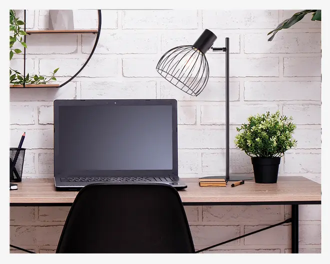 Et moderne arbejdsområde med en slank sort bærbar computer med stor skærm, en minimalistisk sort skrivebordslampe med trådramme og synlig pære, en lille grøn potteplante i sort potte samt et par lukkede notesbøger og en kuglepen på et lyst træskrivebord. Baggrunden er en hvid murstensvæg, og stilen er enkel og nutidig.