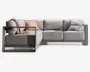 Moderne L-formet sofa med lysegråt betræk og et robust, minimalistisk stel. Sofaen har brede, dybe sæder og tykke ryghynder for ekstra komfort. Der medfølger pynteelementer: et lyserødt plaid, en lyserød pude med frynser og en lysegrå pude samt en åben bog placeret på sædet. Armlæn og bund er lavet af et stilrent materiale med mat finish, hvilket giver sofaen et moderne udtryk.