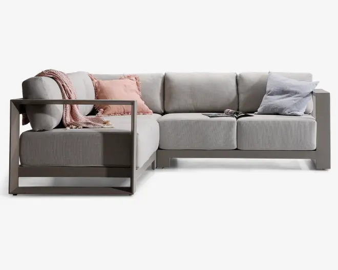 Moderne L-formet sofa med lysegråt betræk og et robust, minimalistisk stel. Sofaen har brede, dybe sæder og tykke ryghynder for ekstra komfort. Der medfølger pynteelementer: et lyserødt plaid, en lyserød pude med frynser og en lysegrå pude samt en åben bog placeret på sædet. Armlæn og bund er lavet af et stilrent materiale med mat finish, hvilket giver sofaen et moderne udtryk.