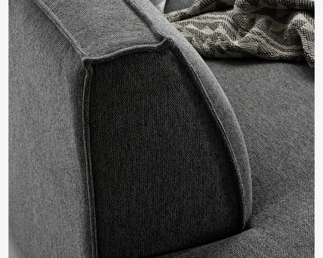 Nærbillede af en sofa-armlæn med tekstureret mørkegrå polstring. Syningerne er tydelige langs kanterne og giver et skræddersyet udtryk. Armlænet er tykt og let afrundet, hvilket giver behagelig støtte. Et mønstret tæppe ligger over sædet og tilføjer kontrast og hygge.