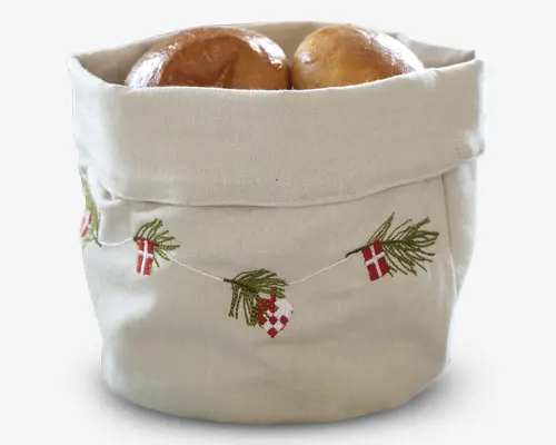 En stofkurv lavet af naturfarvet tekstil med en ombukket kant. Udvendigt er der broderede julemotiver, herunder små røde og hvide flag, grønne grangrene og flettede julehjerter, som hænger sammen som en guirlande. Kurven bruges her til at opbevare eller servere brød.