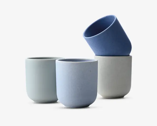 Sæt med fire cylindriske kopper i bløde pastelfarver af blå og offwhite. Hver kop har en glat, mat overflade og et minimalistisk design med lige sider og afrundet bund. Kopperne er uden hank og kan stables, hvilket gør dem velegnede til både varme og kolde drikke. Farverne inkluderer lyseblå, bleg blå, lysegrå og en dybere blå, hvilket giver et moderne og roligt udtryk.