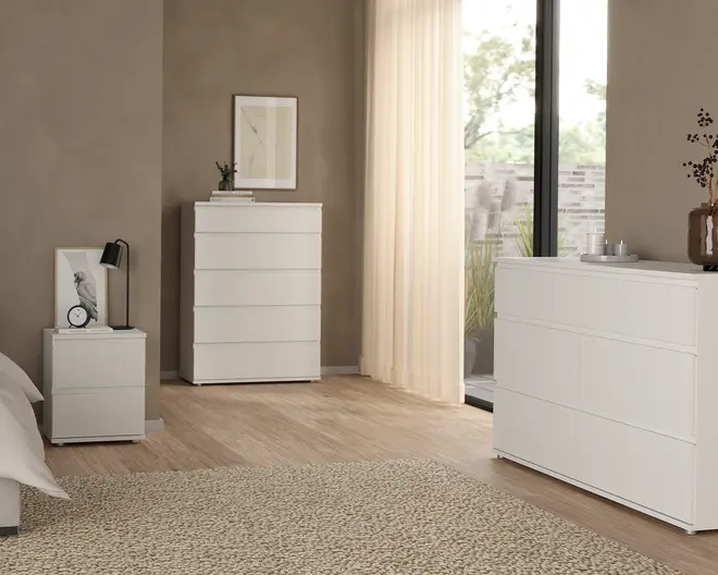 Billedet viser et sæt moderne, minimalistiske hvide opbevaringsmøbler med en glat, mat overflade. Møblerne består af en høj kommode med fem skuffer, en bred kommode med tre skuffer og et lille natbord med to skuffer. Alle enheder har et grebsfrit design for et stilrent udtryk. Skufferne flugter med rammen og skaber rene linjer. Møblerne står på et lyst trægulv, og rummet er indrettet i neutrale toner med enkle accessories som en lampe, et ur, bøger og en vase.