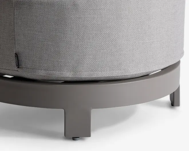 Nærbillede af den nederste del af et rundt møbel med lysegråt, tekstureret stofbetræk. Basen er en solid, glat, mat cirkulær ramme i taupefarve, understøttet af korte, brede ben. Et lille stofmærke ses på syningen af betrækket.
