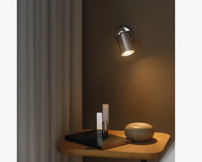 En moderne vægmonteret spotlampe med et slankt, cylindrisk design i metal, der kaster et varmt, fokuseret lys ned på et træbord. På bordet står en rund, glat dekorationsgenstand, to minimalistiske lysestager og et par stablede sorte bøger. Baggrunden har en neutral vægfarve og lyse gardiner, hvilket skaber en hyggelig og moderne stemning.