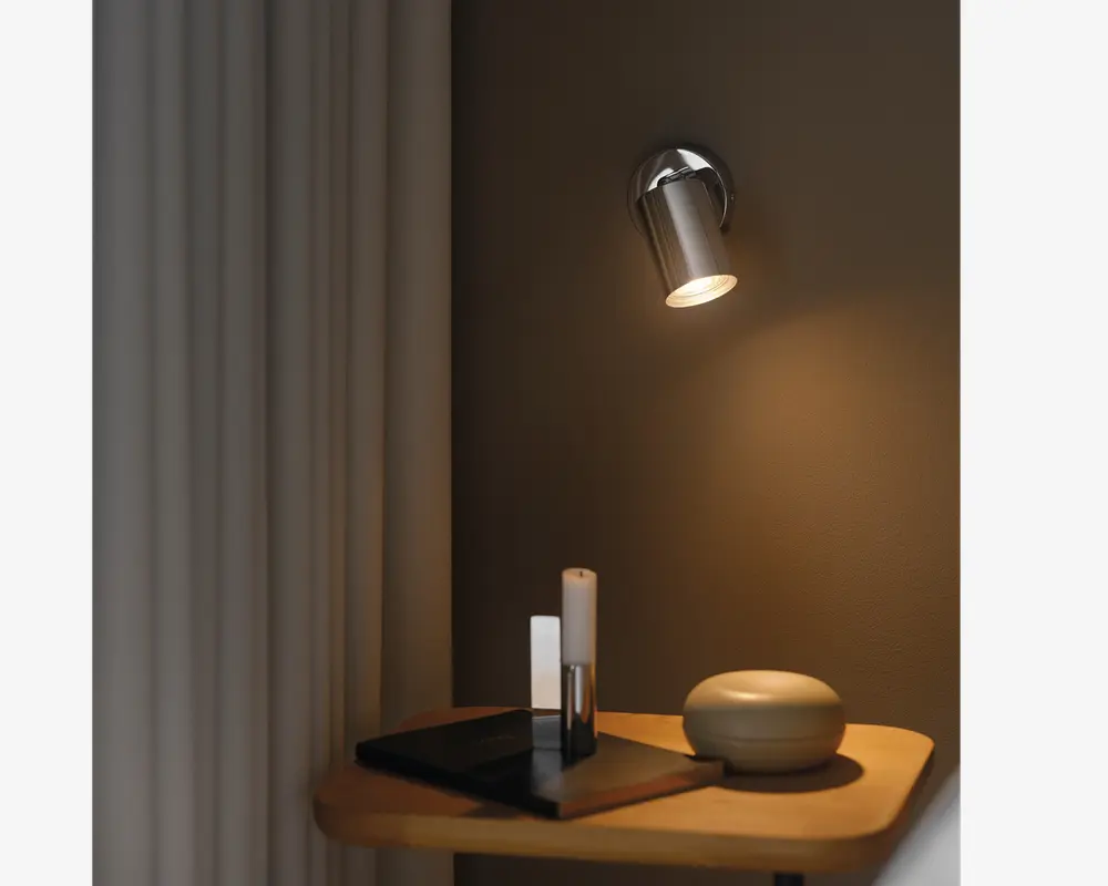En moderne vægmonteret spotlampe med et slankt, cylindrisk design i metal, der kaster et varmt, fokuseret lys ned på et træbord. På bordet står en rund, glat dekorationsgenstand, to minimalistiske lysestager og et par stablede sorte bøger. Baggrunden har en neutral vægfarve og lyse gardiner, hvilket skaber en hyggelig og moderne stemning.
