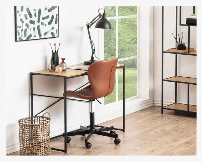 Et moderne hjemmekontor med en brun kontorstol i kunstlæder, der har buet ryglæn og sort metalbase med fem hjul. Stolen står ved et minimalistisk skrivebord med lys træbordplade og sort metalstel. Arbejdspladsen er udstyret med en sort justerbar skrivebordslampe, dekorative genstande og skriveredskaber. Rummet er lyst med naturligt lys fra store vinduer, og gulvet er i lyst træ. En flettet kurv står ved siden af skrivebordet.