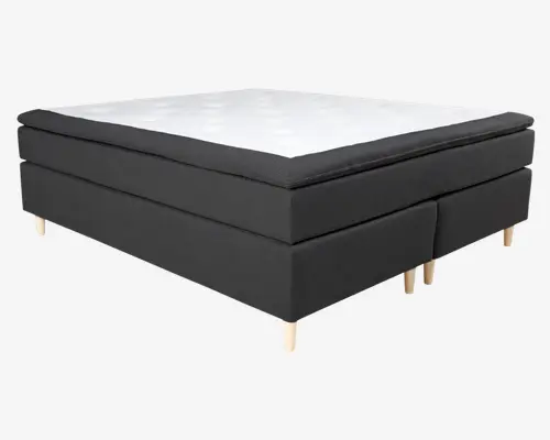 Boxmadras med moderne design, der har en tyk, polstret top i hvid med diskrete syninger for ekstra komfort. Sider og bund er betrukket med mørkt stof, hvilket giver en flot kontrast. Sengen står på korte, lyse træben, som giver et solidt og hævet udtryk. Udtrykket er minimalistisk og tidssvarende, velegnet til forskellige soveværelsesstile.