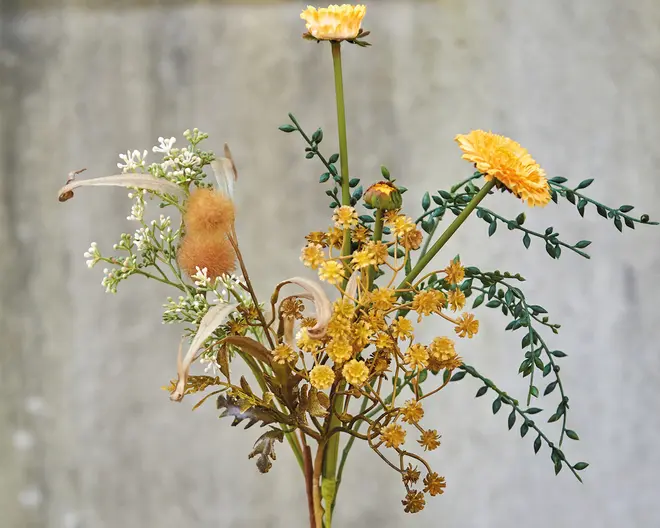 En buket med en blanding af kunstige vilde blomster i gule og hvide nuancer. Arrangementet indeholder små gule blomster, fine hvide blomster og grønne, bladfyldte stilke. Nogle elementer har en lodden, tekstureret overflade, der ligner tørrede græsser. Stilken er slanke, og det samlede udtryk er naturligt og rustikt, velegnet til dekoration.