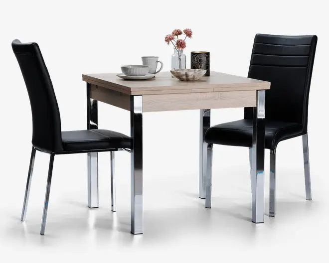 Et kompakt spisesæt bestående af et kvadratisk bord i lyst træ med slanke kromben og to matchende stole. Stolene har sort kunstlæderpolstring med vandrette syninger og kromben. Bordet er dækket med en skål, en kop, en tallerken, en vase med lyserøde blomster og et dekorativt lys, hvilket giver et moderne og indbydende udtryk, der passer til små rum.