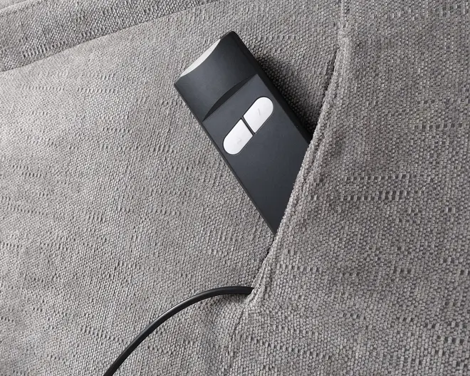 Nærbillede af en grå stoflomme, der er integreret i polstrede møbler, og som indeholder en sort fjernbetjening med ledning og to store hvide knapper. Fjernbetjeningen er delvist synlig, og stoffet har en diskret tekstureret overflade.