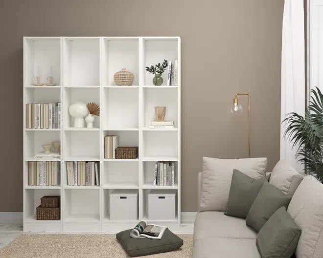 En hvid reol med åbne, kvadratiske rum, der er pænt organiseret med en blanding af bøger, dekorative genstande, flettede kurve og to hvide opbevaringskasser. Nogle hylder viser vaser, en lille plante og skulpturelt pynt, mens andre er fyldt med bøger, der er arrangeret både lodret og vandret. Reolen står op ad en taupefarvet væg og bidrager til et moderne og ryddeligt stue-miljø.