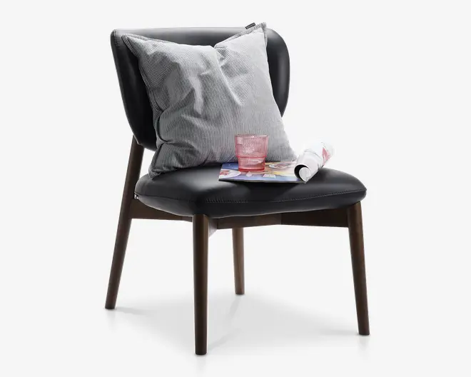 Moderne stol uden armlæn med sort polstret sæde og ryglæn, understøttet af fire ben i træ. Stolen har en stor, lysegrå stribet pude for ekstra komfort. På sædet ligger et lyserødt glas, et sammenrullet magasin og et åbent magasin, hvilket giver et indtryk af en hyggelig og afslappet stemning.
