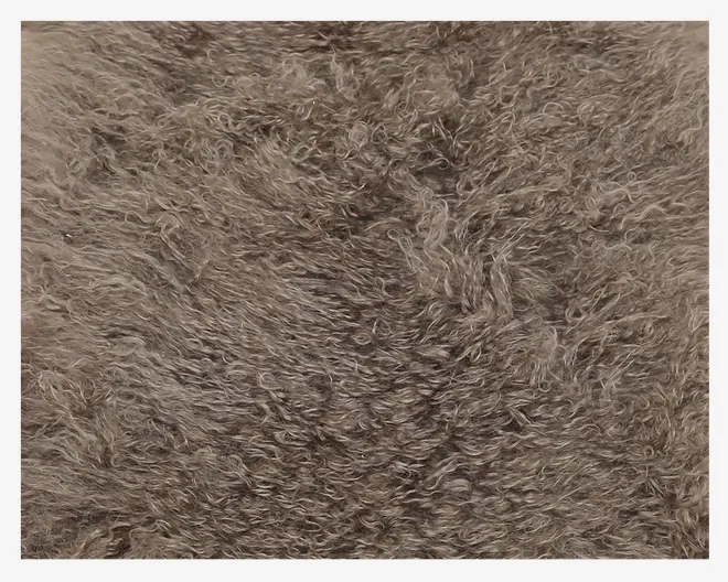 Nærbillede af et blødt, krøllet tekstil i en taupe- eller lysebrun farve. Materialet fremstår fyldigt og tæt, med tætte, snoede fibre, der giver det et luftigt og hyggeligt udtryk, velegnet til varme og komfort.