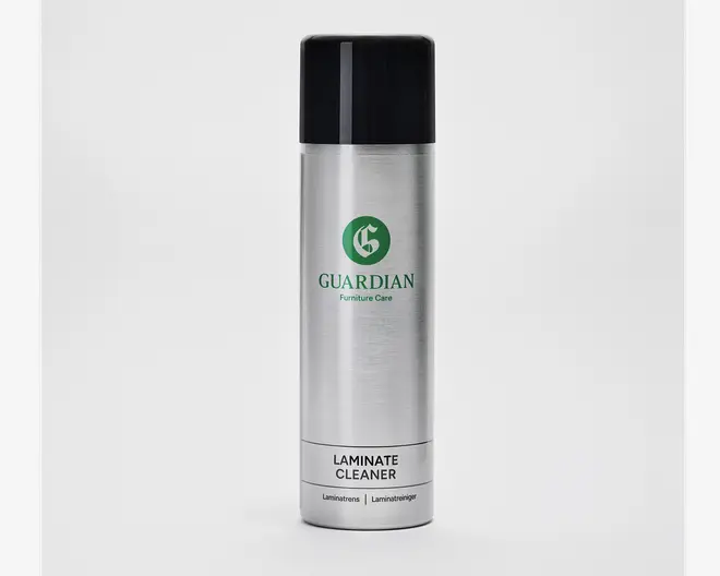 En høj, cylindrisk sølvfarvet spraydåse med sort låg. Dåsen har et grønt logo og mærket 'Guardian Furniture Care' trykt på forsiden. Nederst på etiketten står der 'Laminate Cleaner' med fed sort skrift, og der er yderligere tekst med mindre skrift nedenunder. Designet er stilrent og moderne med børstet metalfinish.