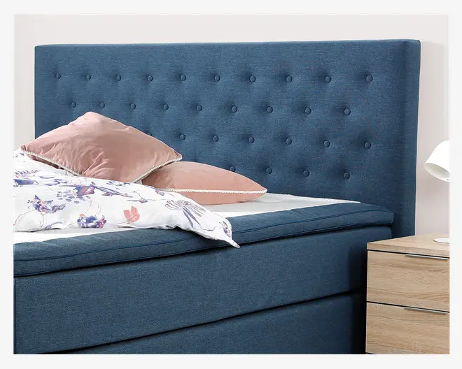 Polstret seng med høj, dybblå stofgavl, der har et knap-tuftet design. Sengen har en tyk, polstret madras og en matchende blå bund. To pudderrosa velourpuder med hvid kant ligger på sengen sammen med et hvidt dynebetræk med lilla og lyserøde blomster. Et natbord i lyst træ med to skuffer og en hvid lampe er delvist synligt ved siden af sengen.