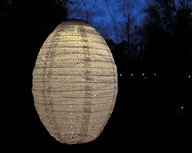 Ovalformet udendørs lanterne med en tekstureret overflade, der har små perforerede huller arrangeret i vandrette rækker. Lanternen udsender et varmt, diffust lys og er ophængt med en tynd wire. Den neutrale farve og det diskrete mønster gør den velegnet til have- eller terrasseindretning og giver stemningsfuld belysning.