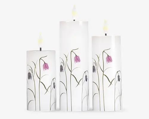 Sæt med tre cylindriske stearinlys i forskellige højder, hver med en glat, hvid overflade. Lysene er dekoreret med et fint blomsterdesign med slanke grønne stilke og lilla, klokkeformede blomster. Vægerne er ubrændte, og det samlede udtryk er elegant og moderne, velegnet til dekoration.