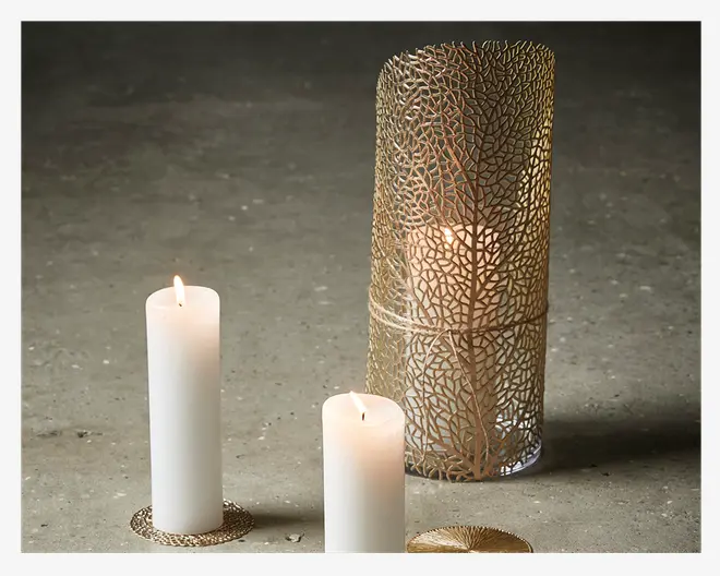 Tre hvide bloklys vises, to står på dekorative, guldfarvede runde underlag med et solstrålemønster, og det tredje er placeret i en høj, cylindrisk holder af guldmetal med et indviklet, bladlignende mønster. Metalholderen giver et elegant, tekstureret udtryk og lader lyset fra lyset skinne igennem mønsteret.