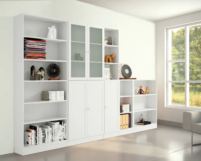 En stor, moderne reol i mat hvid finish med en kombination af åbne hylder, lukkede skabe med solide låger og skabe med matterede glaslåger. Reolen er fyldt med forskellige genstande såsom bøger, magasiner, dekorative objekter og opbevaringskasser. Hylderne er justerbare, hvilket giver fleksibel opbevaring af ting i forskellige højder. Designet er minimalistisk og rent, velegnet til en moderne stue eller et kontor.