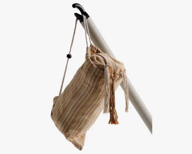Nærbillede af en stofpose med snørelukning, der har et beige og lysebrunt stribet mønster. Posen hænger fra en hvid stang ved hjælp af en justerbar rem med et sort plastspænde. Snoren er bundet i en knude, og posen har en blød, fleksibel form, der egner sig til at bære lette genstande.