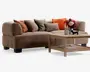 En stor brun, tekstureret sofa med afrundede armlæn og flere pyntepuder i rust, beige, olivengrøn og orange, vist sammen med et lyst sofabord i træ med nederste hylde samt dekorative lys og blomster på toppen.