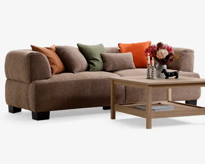 En stor brun, tekstureret sofa med afrundede armlæn og flere pyntepuder i rust, beige, olivengrøn og orange, vist sammen med et lyst sofabord i træ med nederste hylde samt dekorative lys og blomster på toppen.