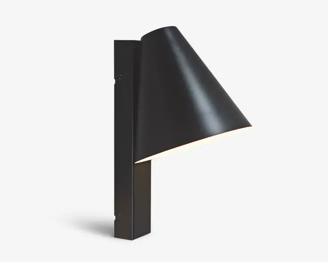 En moderne vægmonteret lampe med mat sort finish. Lampen har en slank, konisk skærm, der leder lyset nedad, og er fastgjort til en rektangulær, lodret base. Designet er minimalistisk og nutidigt, velegnet til både indendørs og udendørs brug. Beslaget er fastgjort med synlige skruer på basen, hvilket understreger det industrielle udtryk.