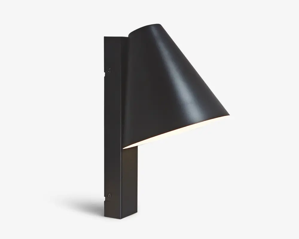 En moderne vægmonteret lampe med mat sort finish. Lampen har en slank, konisk skærm, der leder lyset nedad, og er fastgjort til en rektangulær, lodret base. Designet er minimalistisk og nutidigt, velegnet til både indendørs og udendørs brug. Beslaget er fastgjort med synlige skruer på basen, hvilket understreger det industrielle udtryk.