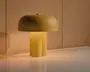 En sennepsgul bordlampe med rund, svampeformet skærm og en skulpturel cylindrisk base, der lyser varmt på en blank overflade foran en lys trævæg.