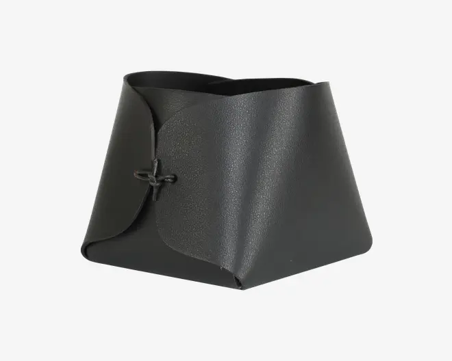 En sort, tekstureret, trapezformet beholder lavet af et enkelt stykke materiale, der er foldet og samlet foran med en simpel knude. Kanterne er glatte, og strukturen er robust med et moderne, minimalistisk design. Åbningen foroven er bred, hvilket gør det nemt at få adgang til indholdet.