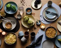 Et træbord dækket med forskellige keramiktallerkener, skåle og kopper i afdæmpede blå, beige og grå nuancer. Service er arrangeret med forskellige madvarer, herunder grønne peberfrugter, artiskokker, sorte druer, pastaskruer, kartoffelchips, peanuts og en pære. Der er også småkager, et glas vand og flere tændte stearinlys. Bestik er placeret på nogle tallerkener, og servietter er foldet ved siden af retterne. Kaffebønner er drysset ud over bordet som dekoration.