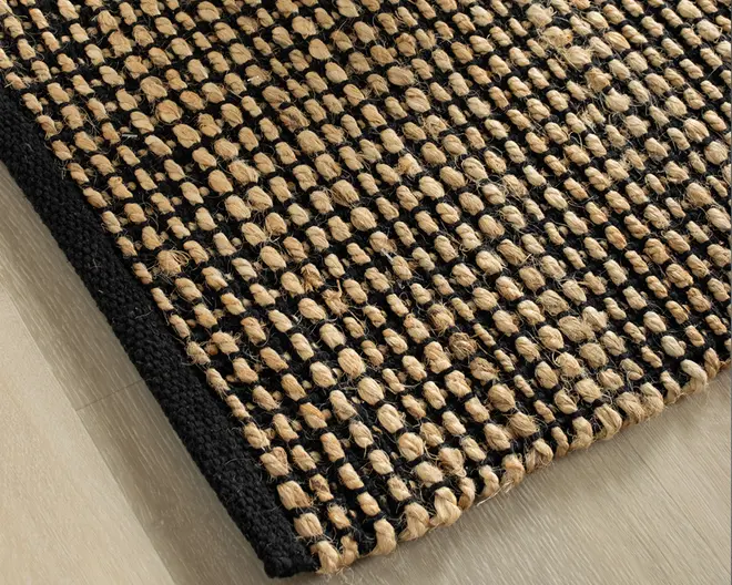 Nærbillede af et vævet tæppe med en tekstureret overflade, hvor tykke, naturlige jute-fibre er flettet sammen med sorte tråde. Tæppet har en kraftig, taktil struktur og en solid sort kant, hvilket giver et moderne og rustikt udtryk. Vævningen danner et gittermønster med hævede, knudrede jute-løkker, der tilføjer dybde og slidstyrke.