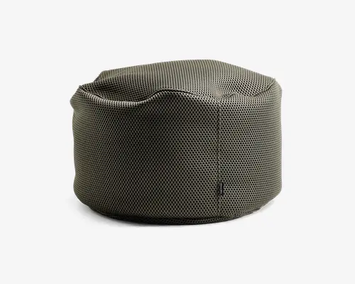 Rund, blød puf med et tekstureret, netlignende stof i en afdæmpet olivengrøn farve. Overfladen har diskrete syningsdetaljer og en let indsunken top for komfortabel siddeplads. Stoffet virker åndbart og slidstærkt, og der ses et lille stofmærke på sidesømmen.