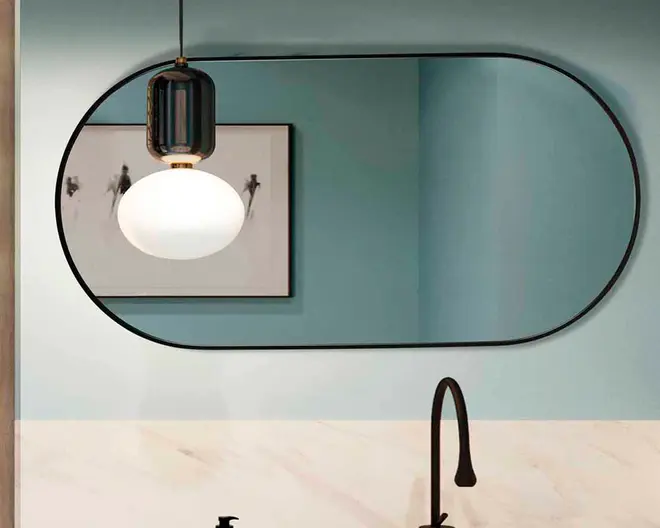 Stort, vandret ovalt spejl med en tynd sort ramme, monteret på en lyseblå væg over en hvid og beige marmoreret overflade. Spejlet reflekterer en moderne pendellampe med sort top og hvid kugle samt et indrammet abstrakt kunstværk. En høj, buet sort vandhane er synlig under spejlet.