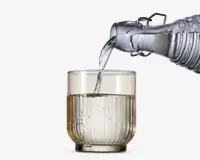 Et klart glas med lodret rillet tekstur fyldes med vand fra en gennemsigtig glaskaraffel. Glasset har en tyk bund og lige sider, og vandet hældes i en jævn strøm.