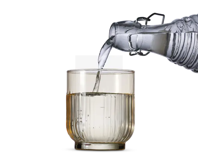 Et klart glas med lodret rillet tekstur fyldes med vand fra en gennemsigtig glaskaraffel. Glasset har en tyk bund og lige sider, og vandet hældes i en jævn strøm.