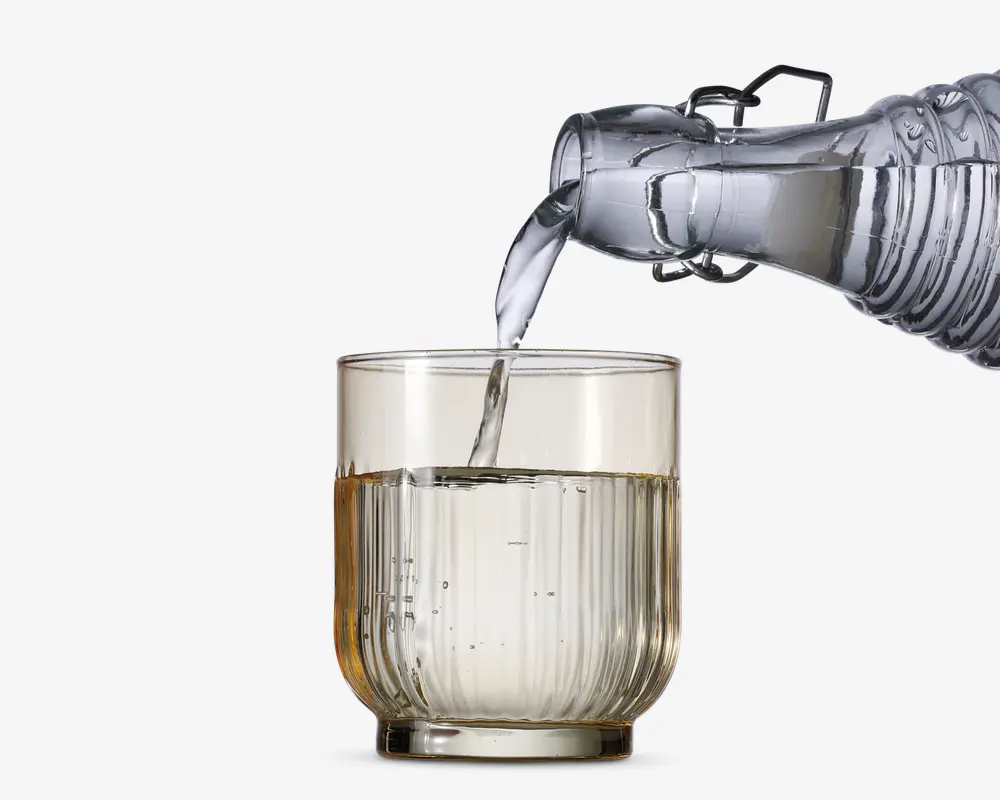 Et klart glas med lodret rillet tekstur fyldes med vand fra en gennemsigtig glaskaraffel. Glasset har en tyk bund og lige sider, og vandet hældes i en jævn strøm.