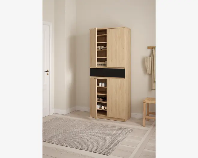 Høj, fritstående skab i lyst træ med naturfinish. Skabet har to lodrette låger, hvoraf den ene er delvist åben og viser flere hylder indeni. Den øverste sektion har hylder til opbevaring af små køkkenapparater og beholdere, mens den nederste sektion har hylder til skåle og andre genstande. Et vandret sort panel adskiller den øverste og nederste sektion og giver et moderne præg til designet. Skabet står på en flad sokkel og har et minimalistisk, moderne udtryk, der passer til køkken eller spisestue.