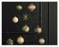 En samling dekorative ornamenter i forskellige former og teksturer, herunder runde, riflede og dråbeformede designs. Ornamenterne har metalliske guld-, grønne og cremefarver med glimmer, prægede mønstre og indviklede detaljer. Nogle har en blank overflade, mens andre er matte eller teksturerede. De hænger på et sort metalstativ med cirkulære holdere, arrangeret i en træ-lignende form mod en mørk væg.