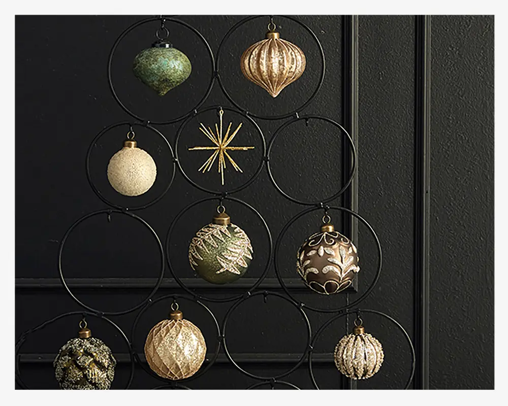 En samling dekorative ornamenter i forskellige former og teksturer, herunder runde, riflede og dråbeformede designs. Ornamenterne har metalliske guld-, grønne og cremefarver med glimmer, prægede mønstre og indviklede detaljer. Nogle har en blank overflade, mens andre er matte eller teksturerede. De hænger på et sort metalstativ med cirkulære holdere, arrangeret i en træ-lignende form mod en mørk væg.