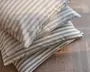 En stak med tre rektangulære puder, hver med et mønster af jævnt fordelte horisontale striber. Striberne er i bløde, neutrale nuancer: den øverste pude har beige striber, den midterste har lyseblå striber, og den nederste har lysegrå striber. Stoffet ser ud til at være en tekstureret bomulds- eller hørblanding, hvilket giver et naturligt og hyggeligt udtryk. Pudernes kanter er pænt syede, og de er fyldige, velegnede til brug på sofaer, stole eller som gulvpuder.
