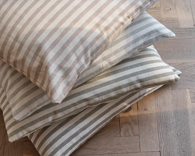 En stak med tre rektangulære puder, hver med et mønster af jævnt fordelte horisontale striber. Striberne er i bløde, neutrale nuancer: den øverste pude har beige striber, den midterste har lyseblå striber, og den nederste har lysegrå striber. Stoffet ser ud til at være en tekstureret bomulds- eller hørblanding, hvilket giver et naturligt og hyggeligt udtryk. Pudernes kanter er pænt syede, og de er fyldige, velegnede til brug på sofaer, stole eller som gulvpuder.