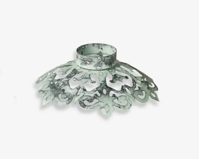 Dekorativt metalstykke med et udsmykket, filigranlignende design og en slidt, antik hvid finish. Produktet har en cirkulær holder øverst, der egner sig til at støtte en cylindrisk genstand, samt en bred, udadbuet base med indviklede udskæringer. Overfladen har et patineret udseende, hvilket giver det et vintage eller shabby chic præg.