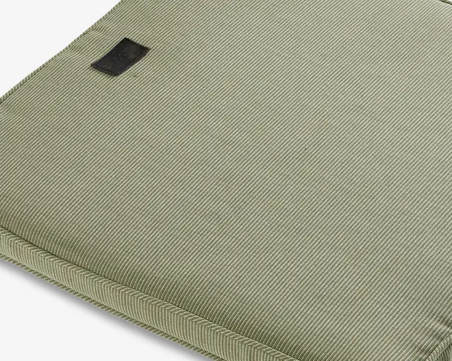Nærbillede af en lyse olivengrøn stofoverflade med en fin, diagonal ribbet struktur. Kanterne er pænt syet, og der er et lille, rektangulært sort mærke syet fast nær det ene hjørne. Materialet virker slidstærkt, og syningerne er præcise, hvilket indikerer en høj kvalitet.